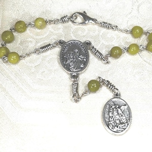 Könnte beinhalten: Ein Rosenkranz mit olivgrünen Perlen und silberfarbenen Metallkomponenten. Der Rosenkranz verfügt über einen Verschluss, der die Perlenkette mit einem zentralen Medaillon und einem ovalen Anhänger mit religiösen Bildern verbindet.