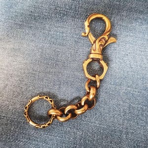 Könnte beinhalten: Ein Schlüsselanhänger aus messingfarbenem Metall mit einem drehbaren Karabinerhaken, der mit einem sechseckigen Glied verbunden ist, gefolgt von einer kurzen Kette aus runden Gliedern und einem Ring mit geprägtem Muster.