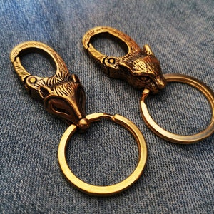 Può includere: Due portachiavi dorati con design a testa di volpe. Ogni portachiavi ha un anello attaccato alla testa di volpe. I portachiavi sono su uno sfondo di jeans blu.