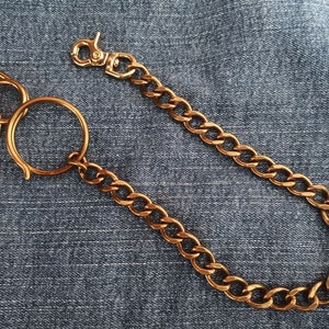 May include: A copper-colored chain with a lobster clasp and a large ring. The chain is made of interlocking oval links. The clasp and ring are also copper-colored. The chain is lying on a blue denim surface.