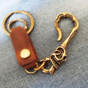 以下が含まれることがあります： 真鍮製のキーホルダー。金色のスナップボタンが付いた茶色のレザーストラップ、円形の二重リング、そして基部に精巧なスカルの装飾が施されたフック型の留め具が、小さな金属製の丸カンで連結されています。