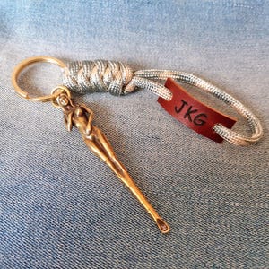 May include: A keychain featuring a gold-toned metal figure of a person, a silver and grey paracord loop with a knot, and a small rectangular brown leather tag embossed with the black letters JKG attached to a gold-toned metal ring.