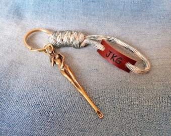 May include: A keychain featuring a gold-toned metal figure of a person, a silver and grey paracord loop with a knot, and a small rectangular brown leather tag embossed with the black letters JKG attached to a gold-toned metal ring.