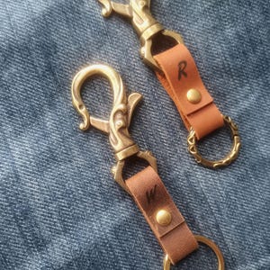 May include: Two keychains featuring antique brass swivel lobster clasps attached to tan leather straps. Each strap is secured with a brass rivet and embossed with a single letter, R and W respectively. A decorative brass ring is attached to the base.