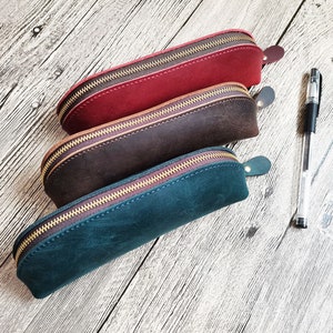 Op de afbeelding: Drie leren etuis in bordeauxrood, bruin en blauwgroen, elk met een gouden rits en een klein leren lipje. Een doorzichtige pen met zwarte inkt ligt naast de etuis op een houten oppervlak.