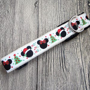 May include: White dog collar with a festive Christmas design. The collar features black Mickey Mouse heads wearing Santa hats, red bows, and green Christmas trees. The word "HO" is repeated. A silver metal D-ring is attached.