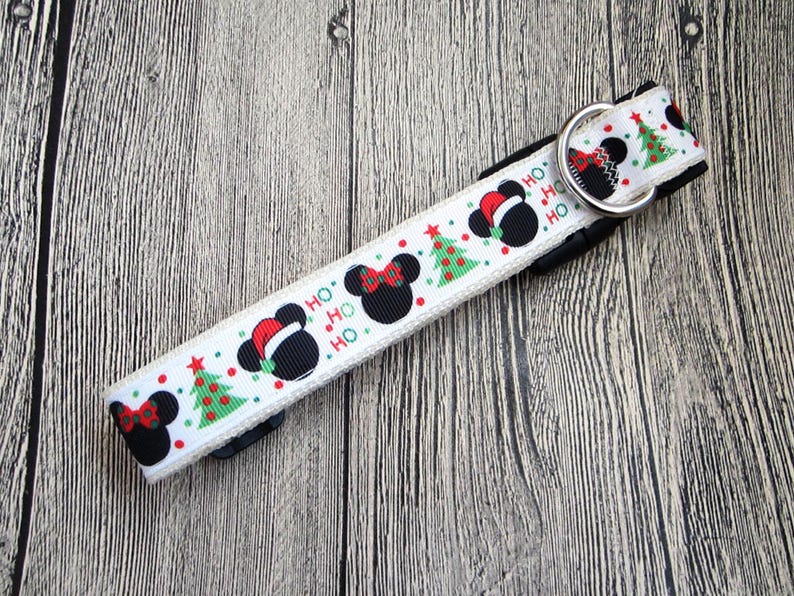 May include: White dog collar with a festive Christmas design. The collar features black Mickey Mouse heads wearing Santa hats, red bows, and green Christmas trees. The word "HO" is repeated. A silver metal D-ring is attached.