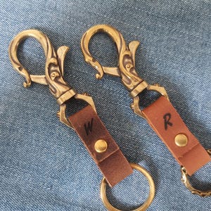 Op de afbeelding: Twee sleutelhangers met sierlijke, draaibare messing karabijnhaken met krulwerk. Elke hanger heeft een bruine leren riem vastgezet met een messing klinknagel, voorzien van de letters W en R, en een ronde messing ring aan de basis.