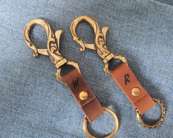 May include: Two keychains featuring ornate brass swivel lobster clasps with scrollwork details. Each has a brown leather strap secured by a brass rivet, with the letters W and R stamped on them, and a circular brass split ring at the base.