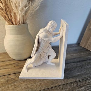 May include: A white stone-textured bookend featuring a sculpted figure of a bearded man in a draped garment, kneeling and pushing against a flat vertical panel. The figure has a muscular build and is depicted in a dynamic, straining pose.