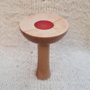 May include: A mushroom-shaped object crafted from light-coloured wood with a smooth, polished finish. The top cap features a circular, vibrant red resin inlay centred on the flat surface, while the stem is cylindrical and tapers slightly outward.
