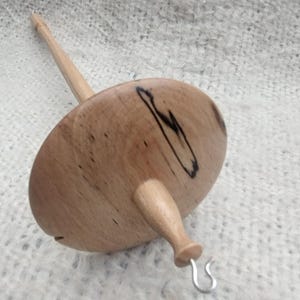 May include: A wooden drop spindle featuring a circular whorl with natural wood grain patterns and dark streaks. The spindle has a long, slender shaft and a metal hook at the bottom, designed for spinning fibre into yarn with a smooth, polished finish.