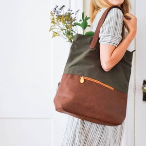 May include: A woman carries a green and brown canvas tote bag with a leather bottom and a zippered pocket. The bag has a gold zipper pull.