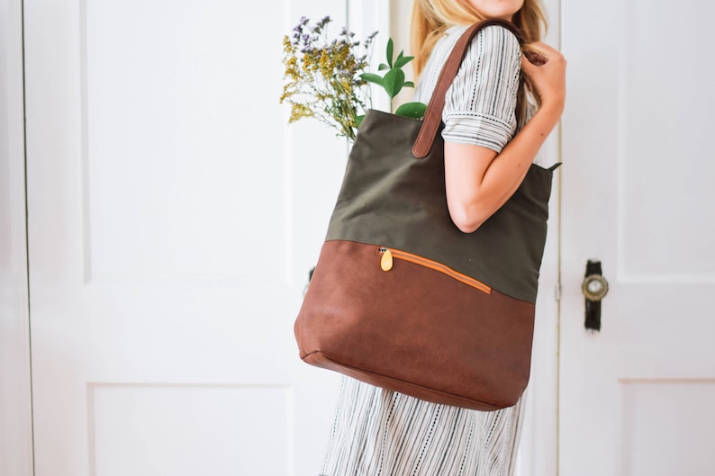 May include: A woman carries a green and brown canvas tote bag with a leather bottom and a zippered pocket. The bag has a gold zipper pull.