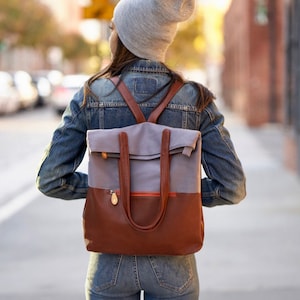 May include: A person wearing a gray beanie and a denim jacket is walking down a city street. They are carrying a brown and gray canvas backpack with leather straps and a flap.