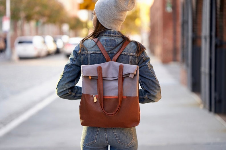 May include: A person wearing a gray beanie and a denim jacket is walking down a city street. They are carrying a brown and gray canvas backpack with leather straps and a flap.