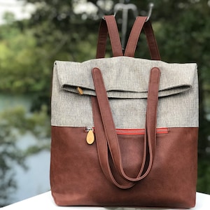 Puede incluir: Una mochila de cuero marrón y lona gris con un bolsillo con cremallera y un cierre de solapa. La mochila tiene dos correas ajustables.