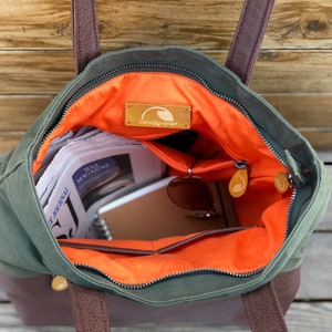 May include: A green canvas tote bag with brown leather handles and a zipper closure. The bag is open and has an orange lining. The bag is filled with items such as newspapers, a notebook, and sunglasses. The bag has a brown leather patch with the text "canopy cove" on it.