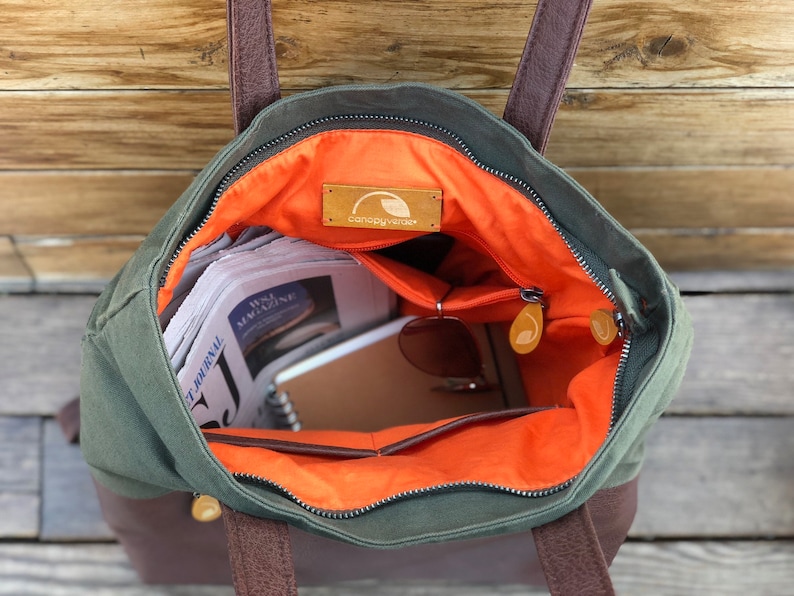 May include: A green canvas tote bag with brown leather handles and a zipper closure. The bag is open and has an orange lining. The bag is filled with items such as newspapers, a notebook, and sunglasses. The bag has a brown leather patch with the text "canopy cove" on it.