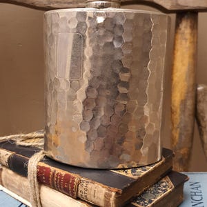 May include: A cylindrical silver-toned metal flask featuring a hammered texture across its surface. The flask has a metallic screw-top cap and a smooth rectangular panel on the front. It is positioned upright on top of two stacked vintage books.