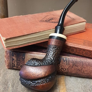 May include: A tobacco pipe featuring a dark brown, textured, rusticated bowl with smooth, polished wooden bands. The pipe has a curved black stem and a cream-colored ring at the junction between the bowl and the stem, resting on two old books.