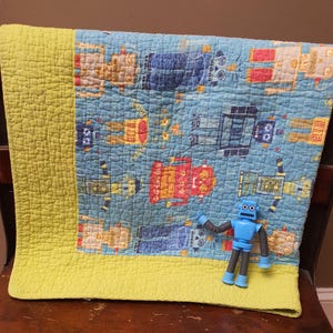 May include: A blue and yellow quilt with a repeating pattern of colorful robots. The quilt is folded over a wooden chair. A small blue robot toy is placed on the quilt.
