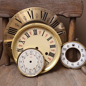 May include: Three circular clock faces of varying sizes. The largest is gold with black Roman numerals. The middle face is cream with black Roman numerals and red fleur-de-lis accents. The smallest is white with black Arabic numerals and floral details.
