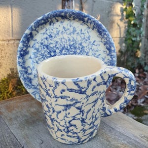 May include: A ceramic mug and saucer set. The mug has a large handle and a cylindrical shape. Both pieces are off-white with a blue splatter pattern. The mug is in front of the saucer, which is slightly out of focus.