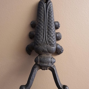 May include: A cast iron boot pull with a dark gray finish. The boot pull has a textured, elongated body with a hole at the top for hanging. Two curved arms extend downward, ending in rounded feet. The design is reminiscent of a stylized insect.