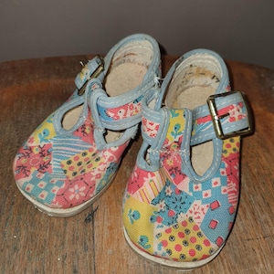May include: A pair of vintage baby shoes with a patchwork design. The shoes feature a light blue trim and straps with gold-colored buckles. The fabric is a mix of floral and geometric patterns in pink, yellow, and blue. The soles are white.
