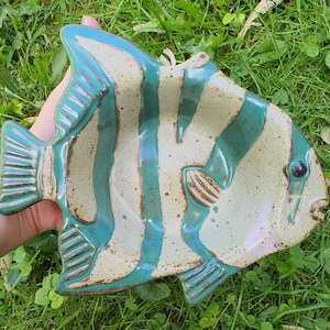 May include: A ceramic fish-shaped serving dish with a teal and off-white color scheme. The dish features teal stripes and fins, with a textured surface. The eye is a small black dot. A unique piece for serving appetizers or as a decorative item.