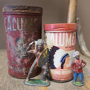 May include: An antique still life featuring two vintage Calumet Baking Powder tins and two small Native American figurines. The tins are red with faded text and graphics. The figurines are painted with vibrant colors.