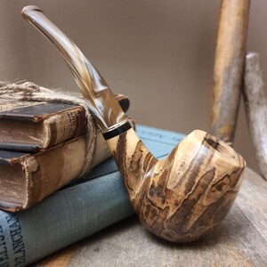 May include: A tobacco pipe featuring a polished wooden bowl with a distinct dark brown and tan marbled grain pattern. The stem is made of a translucent, swirled beige and white acrylic material, connected to the bowl by a thin black and silver ring.