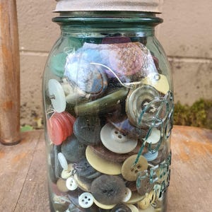 May include: A clear glass jar filled with a variety of colorful buttons. The buttons are in various sizes, shapes, and colors, including white, black, brown, and green. The jar has a metal lid and sits on a wooden surface.