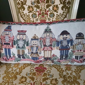 May include: A rectangular throw pillow featuring a woven tapestry design of seven nutcracker figures in various uniforms, including red, green, and blue, accented with holly leaves and a red ribbon border on a cream-colored background.