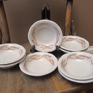 May include: A set of six white ceramic bowls with a brown leaf design around the rim. The bowls are stacked on a wooden surface, with one bowl displayed on a black stand. The bowls have a scalloped edge.