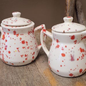 May include: A ceramic sugar bowl with two handles and a matching lid, paired with a ceramic creamer pitcher featuring a lid. Both pieces are off-white with a speckled pattern of red and orange paint splatters across their surfaces.