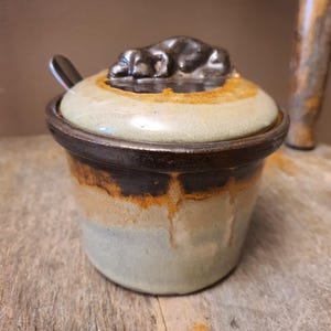 May include: A ceramic lidded jar with a small metal spoon, featuring a multi-toned glaze in shades of cream, tan, and dark brown. The lid has a sculpted, dark brown handle, and the body displays a drip-style glaze pattern with a smooth finish.