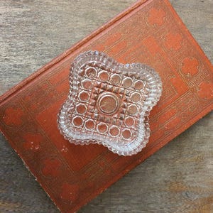 May include: A clear, pressed glass dish with a scalloped, four-lobed edge and a geometric pattern of raised squares and a central circle. The dish features a textured, faceted surface that catches light, creating a repeating grid design.