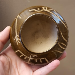 May include: A brown ceramic bowl with a circular shape and a light-colored interior. The exterior features etched white designs, including a bird and geometric shapes. The bowl is held in a hand, showcasing its size and form.