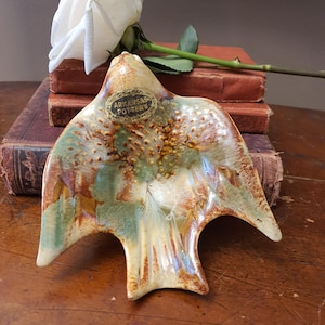 May include: A ceramic bird-shaped dish with a mottled glaze in shades of brown, green, and cream. The dish has a label that reads "ARKANSAS POTTERY". A white rose rests on a stack of old books.