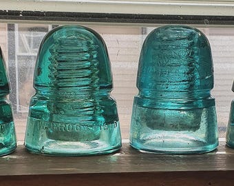 May include: Four antique teal glass insulators with a ribbed design. The insulators are arranged in a row and are made of transparent glass. The base of each insulator has embossed text. The insulators are displayed on a wooden surface.