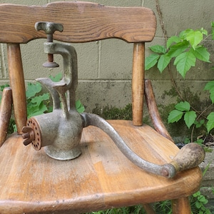 May include: An antique, hand-crank meat grinder made of cast iron, resting on a wooden chair. The grinder has a clamp for securing it to a surface, a handle, and a grinding mechanism. The wood chair has a natural finish.