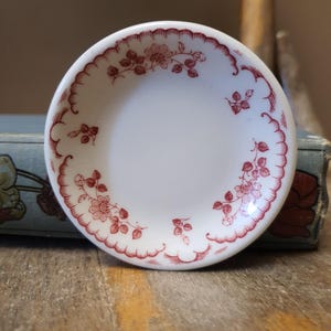 May include: A small, round white ceramic saucer featuring a delicate red floral pattern along the inner rim. The design consists of stylized flowers and leaves arranged in a repeating motif, with a scalloped red line bordering the edge of the dish.