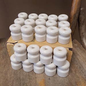 May include: A collection of white ceramic electrical insulators with a spool shape and a central hole, arranged in rows on top of a cardboard box. The box features printed text including the words 'BUILDER' and 'TENTH EDITION' on its side panels.