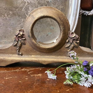 May include: A weathered gold-painted wooden clock casing featuring a central circular opening and decorative floral carvings on each side. The structure has a wide base and visible cracks in the finish, accompanied by a small cluster of white and purple flowers.