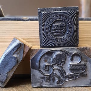 May include: Three vintage metal printing blocks. One block has the text "HIGH GRADE FOOD" in a circular pattern. Another block depicts a person with a bowl. The third block shows a hand. All blocks are mounted on wooden bases.