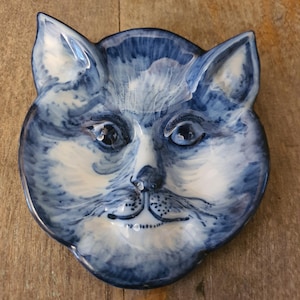 May include: A ceramic trinket dish shaped like a cat head, featuring a blue and white hand-painted finish. The design displays detailed brushstrokes depicting the cat's fur, eyes, nose, and whiskers, with a glossy surface and raised ear edges.
