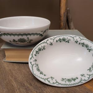 May include: A white ceramic bowl and a matching oval dish, both featuring a decorative green floral pattern along the edges. The bowl is deep with a rounded rim, while the dish is shallow and elongated, showcasing the same intricate botanical design.