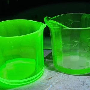 May include: Two vintage green glass pieces. One is a mug with a handle, and the other is a small pitcher with a handle and a decorative etched design. Both pieces have a bright, neon green color.